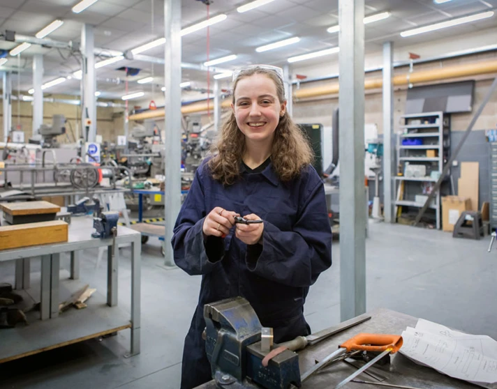 A female mechanical engineering student in navy-blue overalls smiling while working on a small metal component in a busy college workshop. A female mechanical engineering student in navy-blue overalls smiling while working on a small metal component in a busy college workshop.